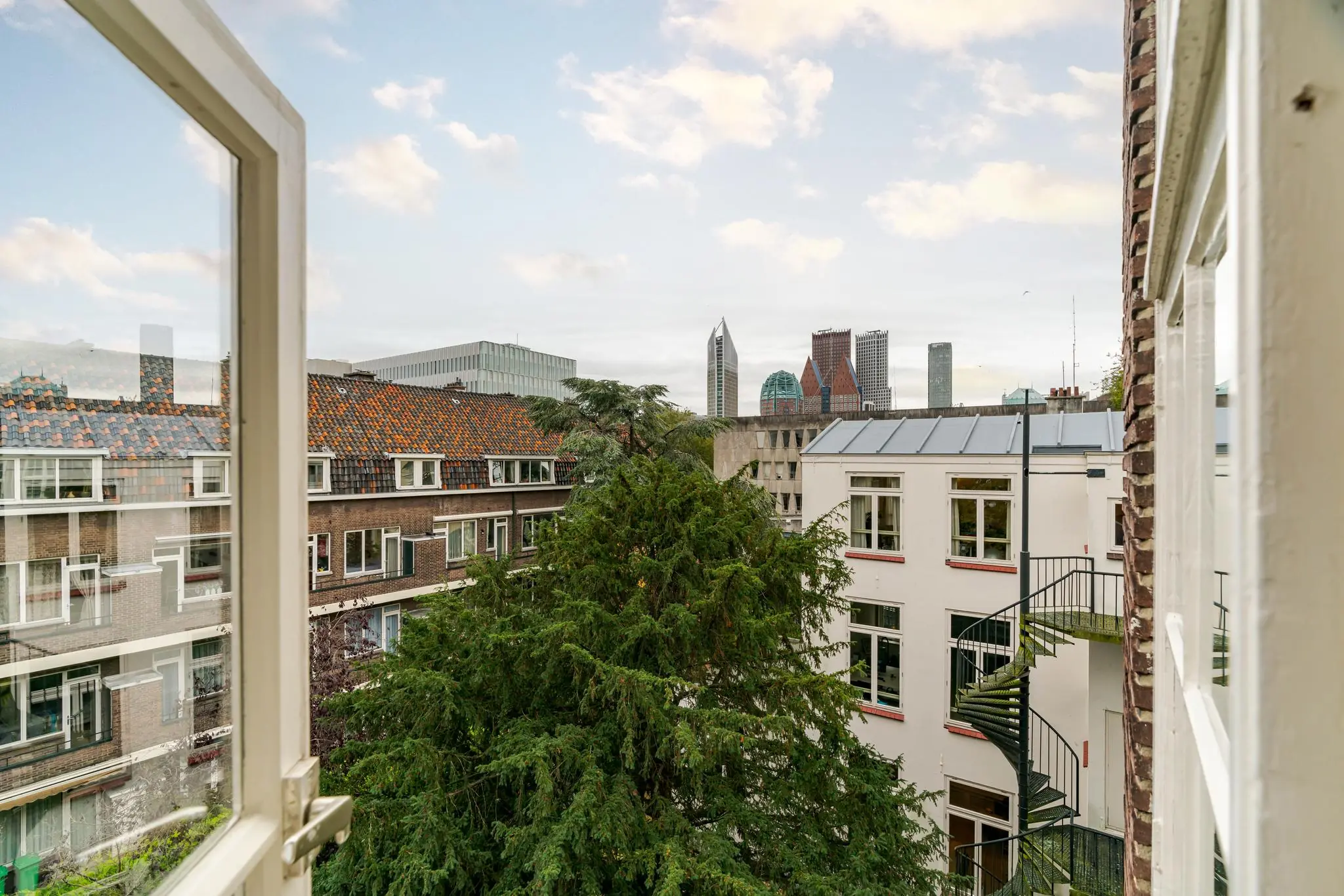 Uitzicht vanuit een open raam op het Lange Voorhout in Den Haag met zicht op diverse stadsgebouwen en wolkenkrabbers op de achtergrond.